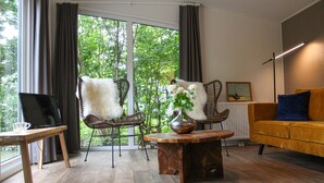 Ein modernes Wohnzimmer mit einem großen Fenster, zwei Korbstühle mit weißen Kissen, einem Holztisch mit Blumenvase, einem kleinen Tisch mit einem Glas und einem gelben Sofa.