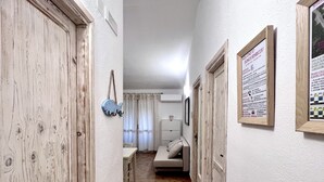A narrow hallway with wooden doors, a white wall, and a tiled floor. There is a bed with a white headboard and a small table with a chair in the background.