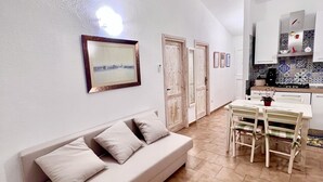 A modern living room with a sofa, dining table, and chairs. There are doors leading to other rooms, and a kitchen area with cabinets and a stove.
