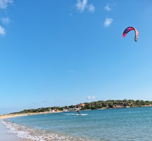 A beach with a clear sky, a person kite surfing, and buildings on a hill in the background.