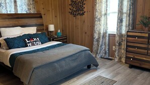 Quarto com cama, ventoinha no teto, paredes de madeira, mesa de cabeceira, penteadeira e janela com cortinas.