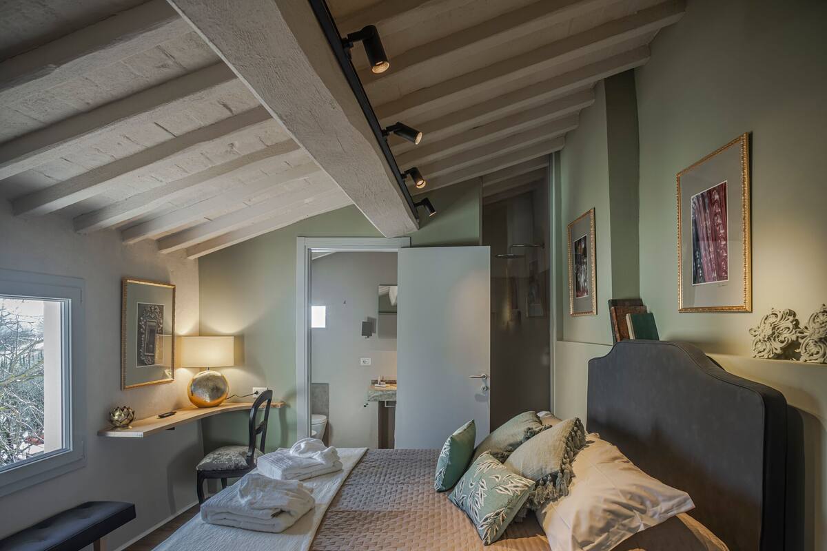 A bedroom with a bed, a bench, a bedside table, a lamp, and a view of the bathroom.