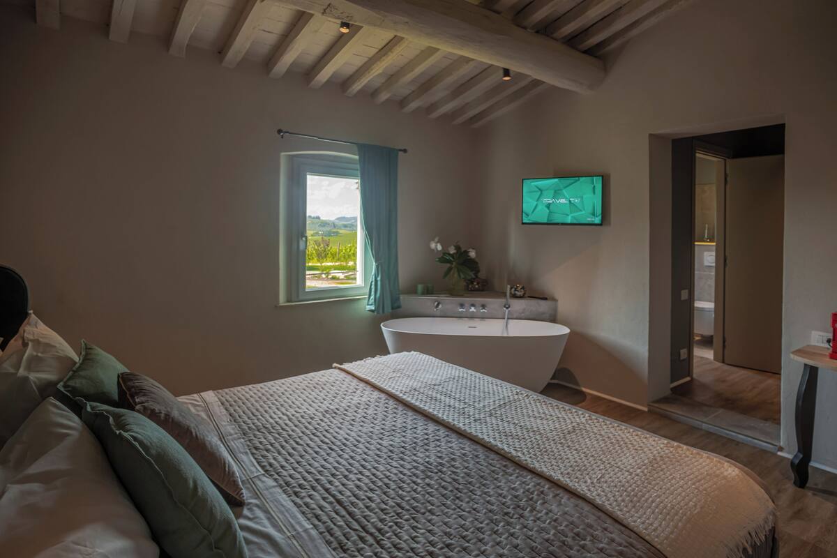 A bedroom with a large bed, a bathtub, a window with curtains, and a TV on the wall.