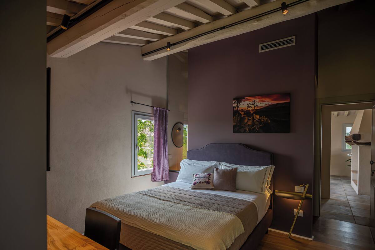 A bedroom with a bed, a window with curtains, a painting on the wall, and a wooden table with a remote control and mug.
