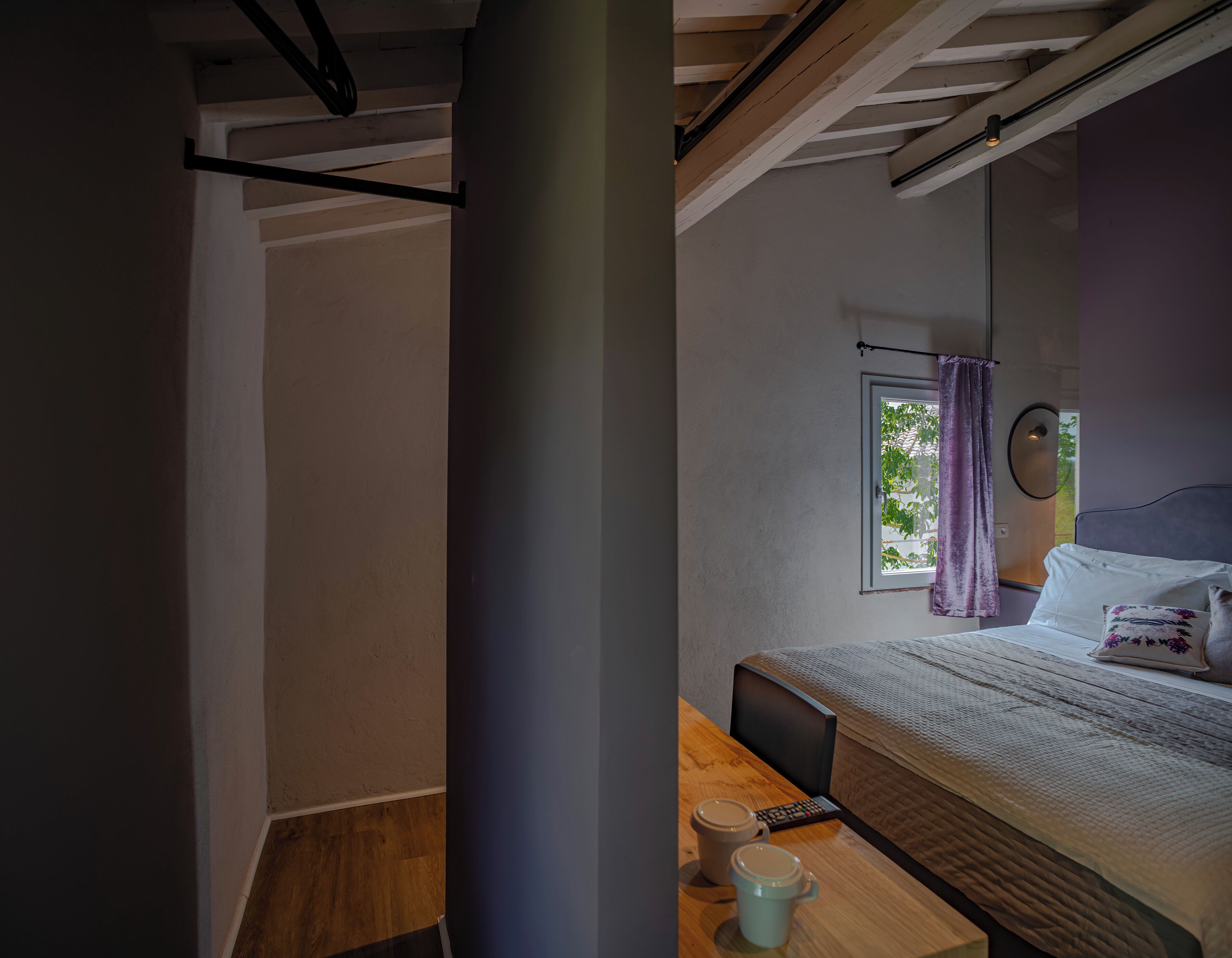 Ein Schlafzimmer mit einem Bett, einem Fenster mit Vorhängen, einem Nachttisch mit Tasse und Fernbedienung sowie einem Holzboden.