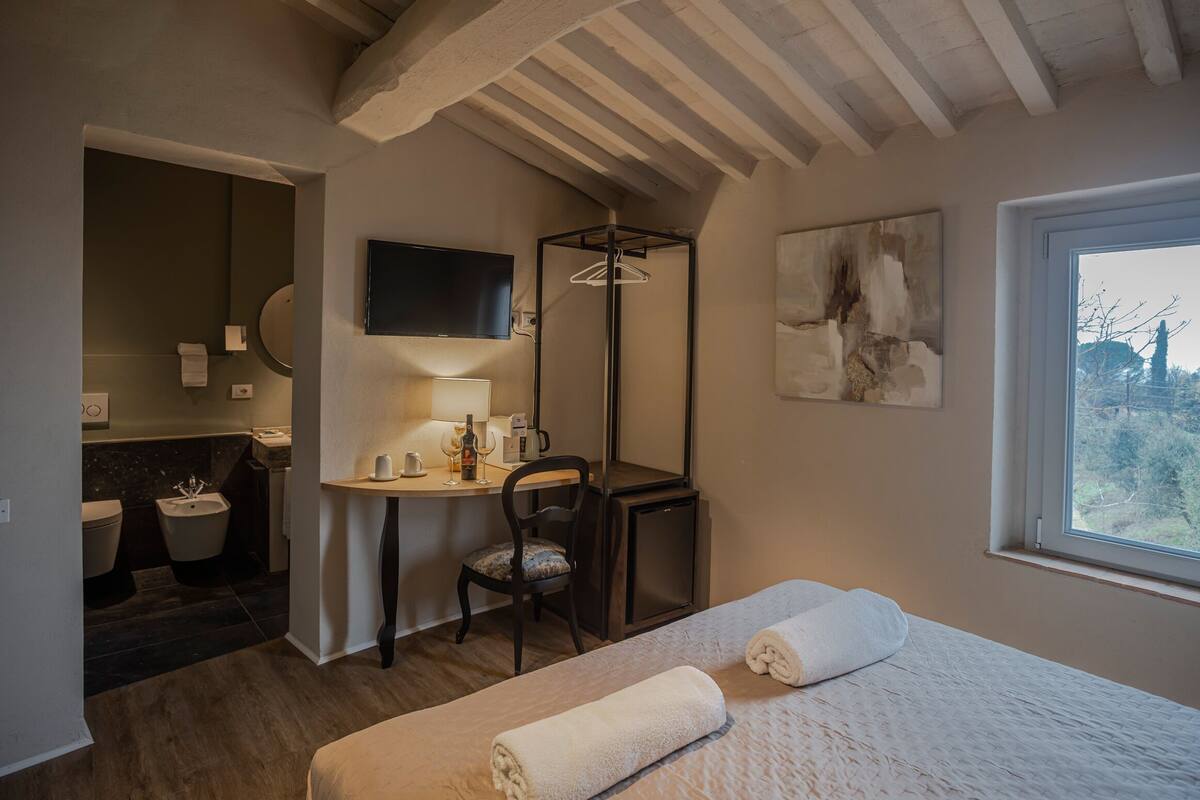 A hotel room with a bed, a desk with a chair, a TV, a window, and a bathroom.