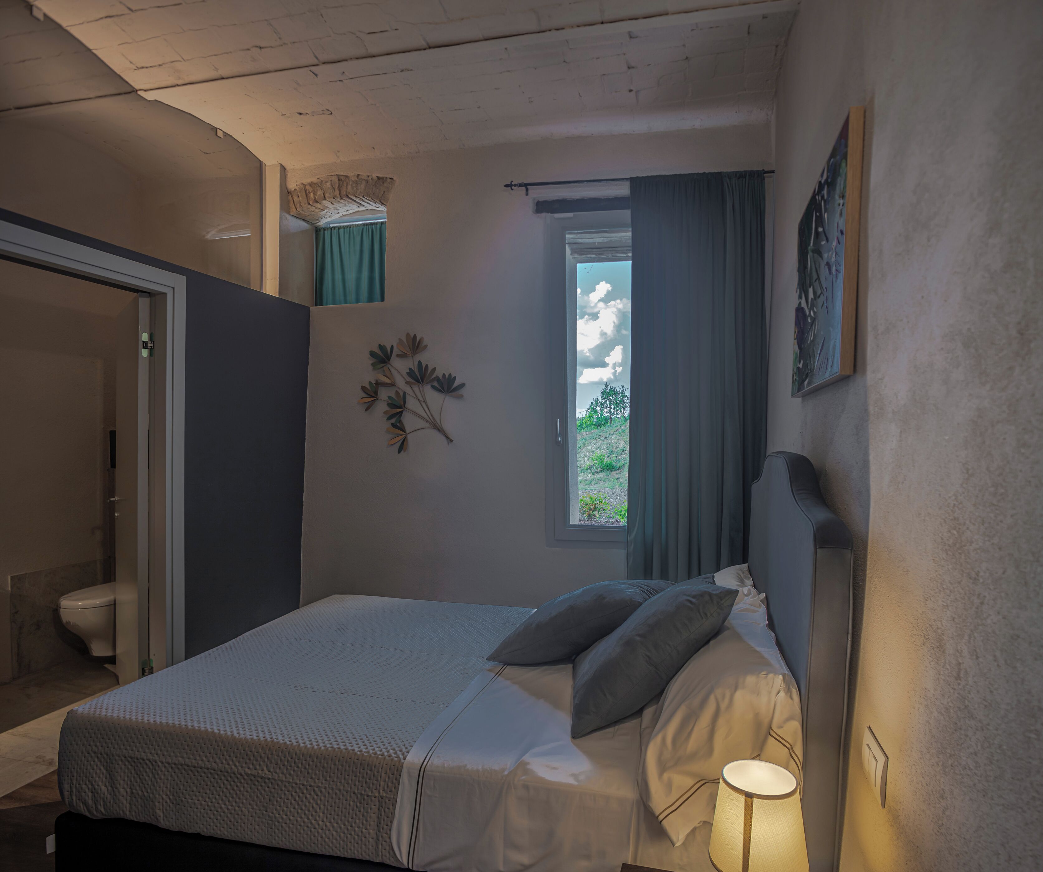Un dormitorio con una cama, una ventana con cortinas, una lámpara de noche y una planta en la pared.