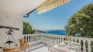 Een balkon met een telescoop, rieten stoelen en een eettafel, met uitzicht op zee.