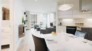 Una cocina moderna con armarios blancos, un comedor con una mesa blanca dispuesta para dos personas y una sala de estar con sofá y sillones.