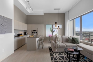 A modern apartment with a kitchen, dining area, and living room. It features a large window with a city view, a sofa with a throw blanket, and a coffee table.