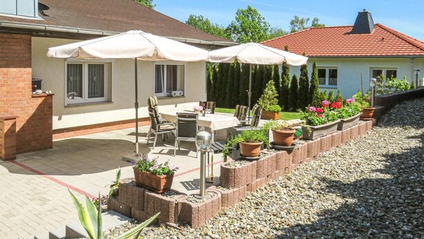 Un patio amueblado con mobiliario exterior y sombrillas, rodeado de plantas en macetas y un muro de ladrillo.