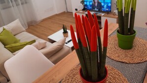 Uma sala de estar moderna com sofá, mesa de centro e televisão exibindo uma transmissão de notícias. Há plantas em vasos sobre a mesa de centro.