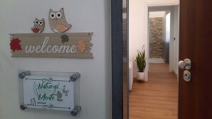 Uma placa de boas-vindas com corujas de madeira e folhas de outono, um letreiro emoldurado com a inscrição 'Natural Mente apartment' e uma porta de madeira que dá para um corredor.