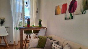 Uma sala de estar com um sofá bege, uma mesa de jantar com cadeiras, uma planta em vaso e uma janela com cortinas.