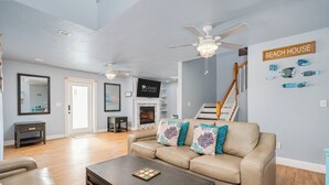 A spacious living room with a beige sofa, a coffee table, and a fireplace. There is a staircase leading to another level, and a sign on the wall that reads "BEACH HOUSE".