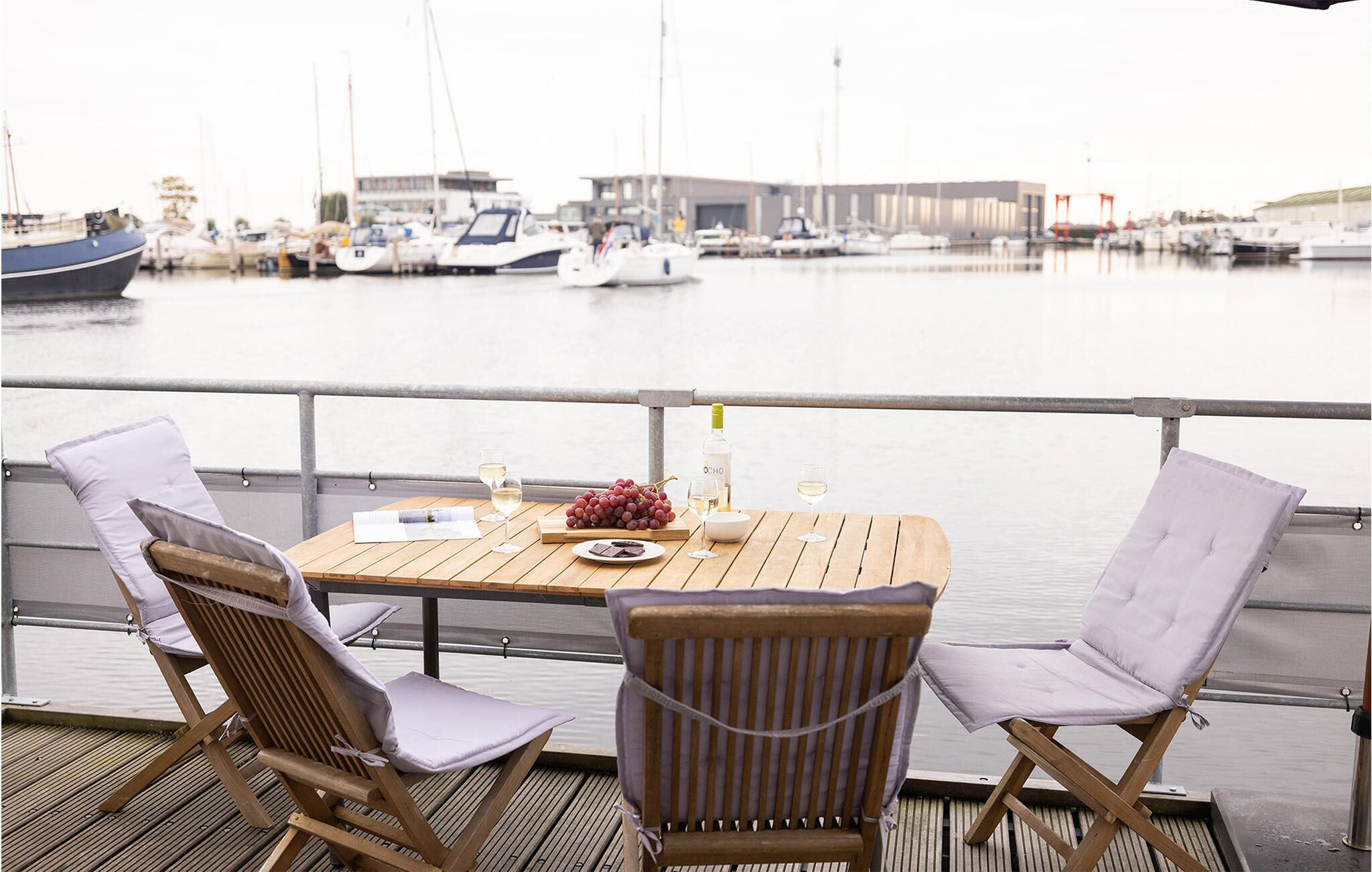 A wooden deck with two cushioned chairs and a table set with a bottle, glasses, and grapes.