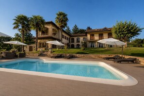 Una piscina amb gandules i ombrel·les, un edifici de pedra amb un balcó i un jardí ben cuidat.