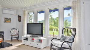 A living room with a flat-screen TV on a white cabinet, a black wicker chair, and a view of greenery through large windows. - Beautiful Home in Torsby With 2 Bedrooms and Wifi (Torsby)