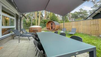An outdoor patio with a long table and chairs, a wooden barrel structure, and a wooden fence in the background.