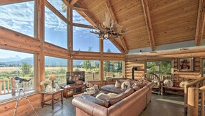 Interior de una cabaña de madera con un amplio sofá de cuero, techo de madera y vista panorámica a las montañas a través de grandes ventanales.