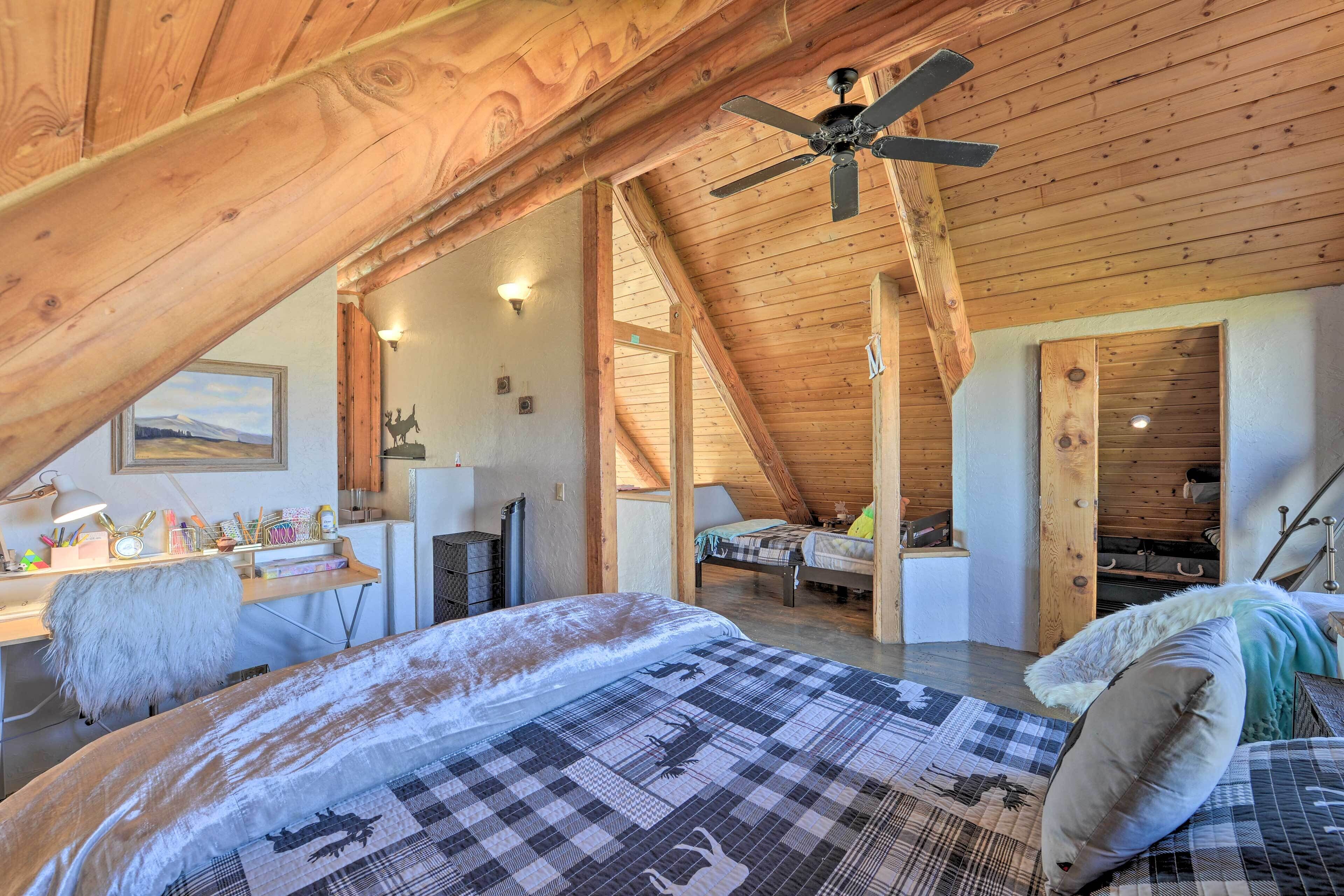 Ein gemütliches Schlafzimmer im Dachgeschoss mit schrägen Wänden, einem Bett mit karierter Bettdecke, einem Schreibtisch mit Lampe und einem Deckenventilator.