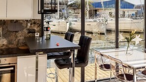 Una cocina moderna con encimera de mármol, comedor con mesa y sillas de vidrio, y vista a barcos a través de grandes ventanales.