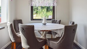 A dining area with a round table, six upholstered chairs, a lace tablecloth, a candle in a glass holder, and a window with a view of greenery.