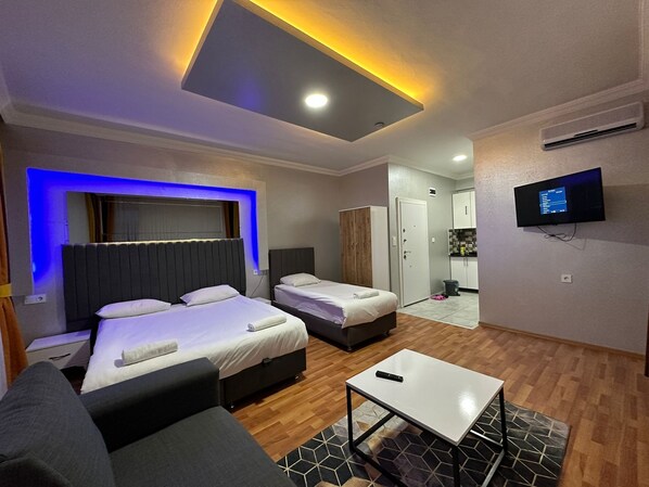 A modern hotel room with two beds, a sofa, a TV, and a small kitchen area.