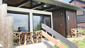 Una terraza de madera con una mesa y sillas de madera, una puerta corrediza y una estructura de techo metálico.