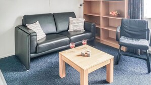 Sala de estar con un sofá de cuero negro, una mesa de centro de madera y un sillón negro. Hay un microondas en un estante y una cesta de fruta en el estante.
