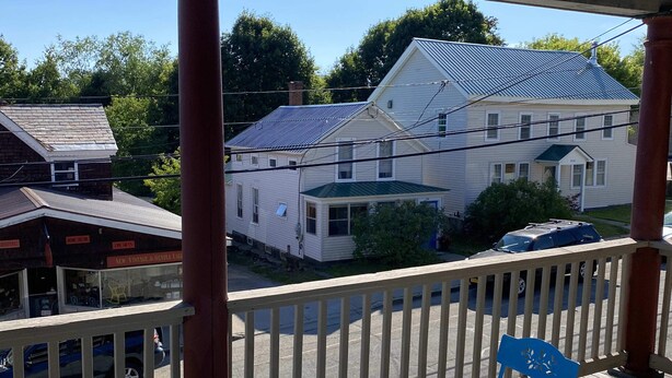 Een overdekt terras met een tafel en stoelen, uitkijkend op een woonwijk met huizen en een straat.