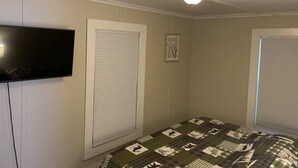 Een slaapkamer met twee ramen, een plafondventilator, een flatscreen-televisie aan de muur en een sprei op het bed.