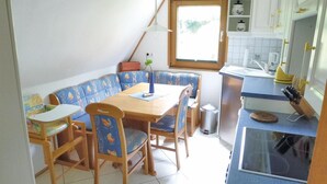 Un coin repas avec une table et des chaises en bois, un banc orné d’un motif floral et une fenêtre donnant sur un paysage verdoyant.