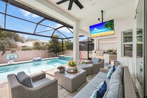 A covered outdoor area with a pool, wicker furniture, a television, and a kitchen area.