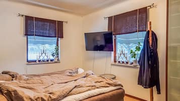A bedroom with a bed, a TV mounted on the wall, and two windows with blinds.