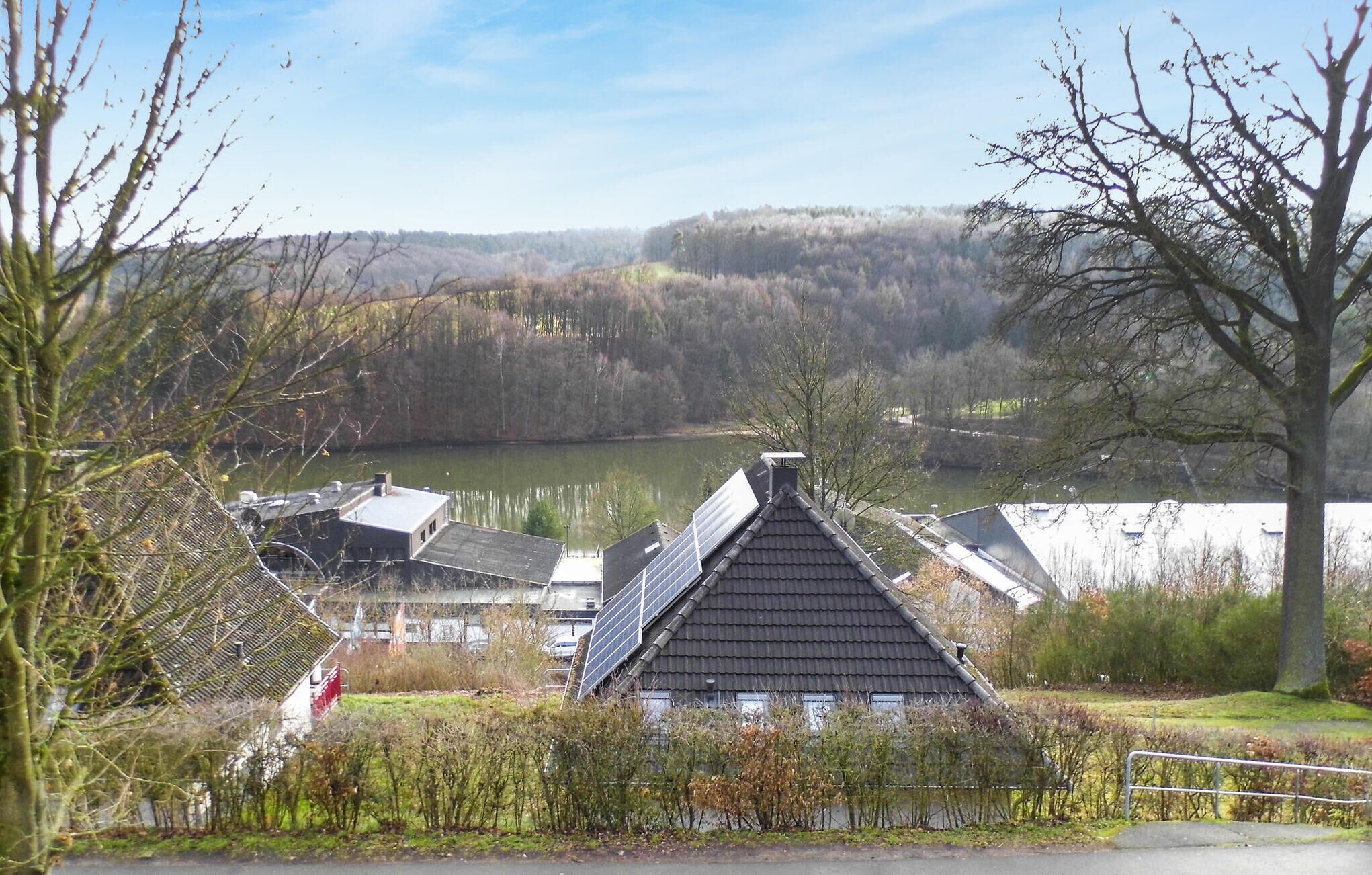 Una casa con techo inclinado, paneles solares y vista a un lago y colinas boscosas al fondo.