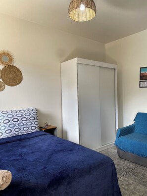 A bedroom with a bed, a wardrobe, a blue chair, and a hanging light fixture. - Apartamento encantador (Florianópolis)