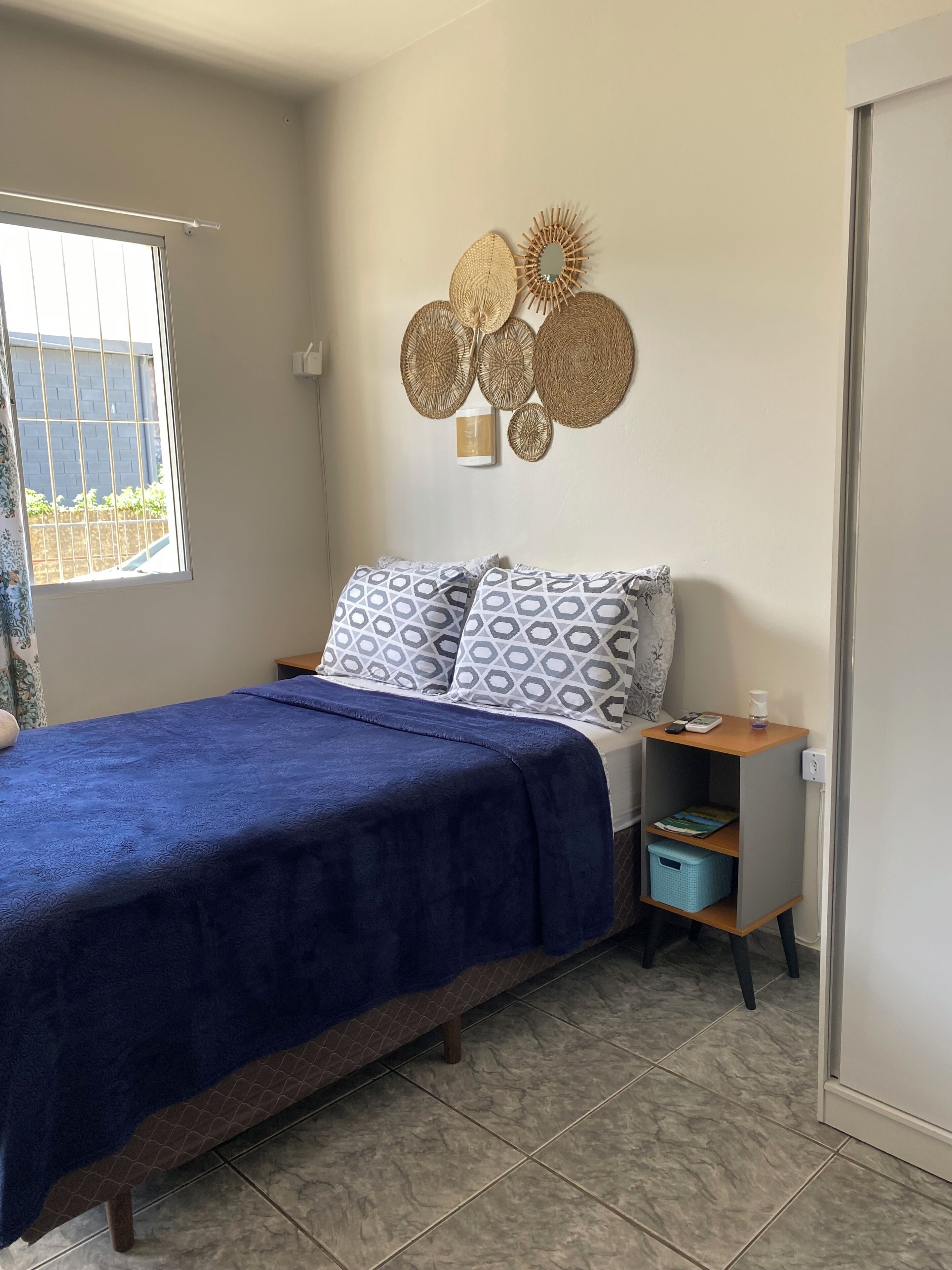 A bedroom with a bed, a bedside table with a lamp, a wall-mounted decorative mirror, and a window with a view of greenery.