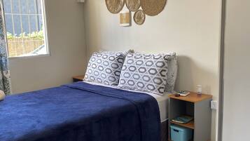 A bedroom with a bed, a bedside table with a lamp, a wall-mounted decorative mirror, and a window with a view of greenery.