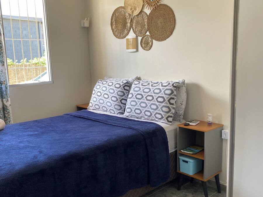 A bedroom with a bed, a bedside table with a lamp, a wall-mounted decorative mirror, and a window with a view of greenery.