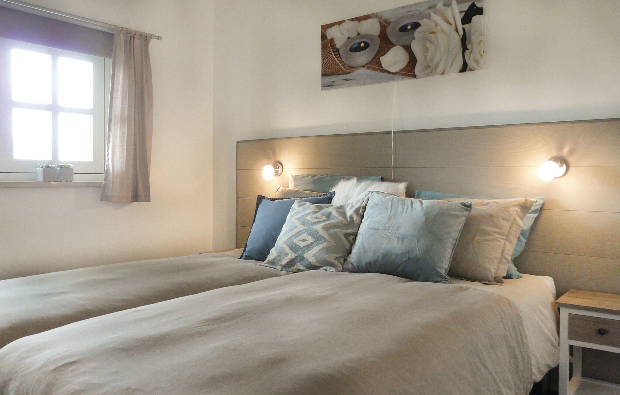 A bedroom with two beds, a headboard with integrated lighting, a bedside table with a drawer, and a window with curtains.