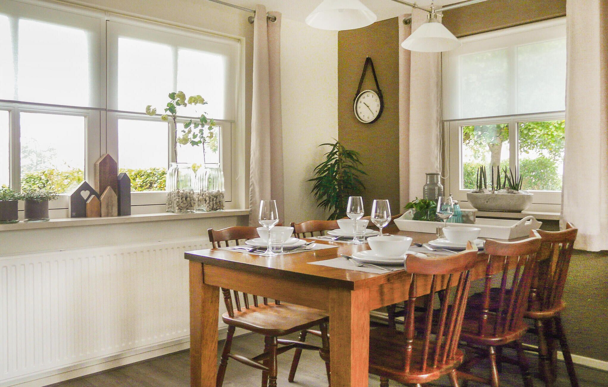 Comedor con una mesa de madera dispuesta para cuatro personas, rodeada de sillas. Hay plantas en macetas en el alféizar de la ventana, un reloj de pared y una ventana con vistas a la vegetación.