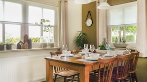 A dining area with a wooden table set for four, surrounded by chairs. There are potted plants on the windowsill, a wall clock, and a window with a view of greenery outside.