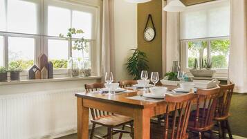 Comedor con una mesa de madera dispuesta para cuatro personas, rodeada de sillas. Hay plantas en macetas en el alféizar de la ventana, un reloj de pared y una ventana con vistas a la vegetación.