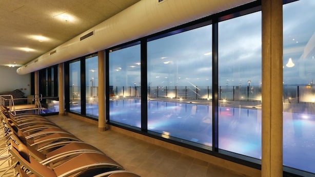 Uma área de piscina com paredes de vidro transparente, espaço de estar com espreguiçadeiras e vista da piscina através do vidro.
