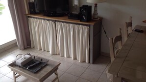 Ein Wohnzimmer mit einem Flachbildfernseher, einer Lampe, einem Tisch mit einem Laptop und einem Essbereich mit Stühlen.