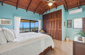 A bedroom with a bed, a ceiling fan, a view of the ocean, a sliding door, and a potted plant.