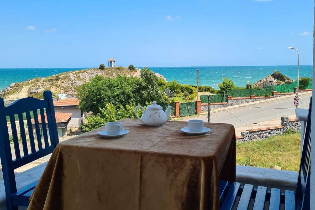 A table with a tablecloth, two chairs, and a teapot set on a balcony overlooking a coastal view.