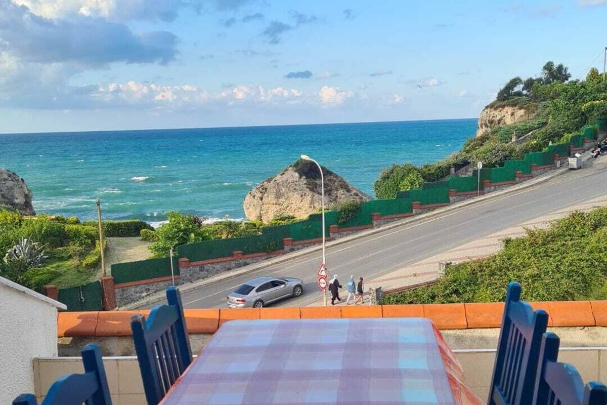 A table with a checkered tablecloth and blue chairs on a balcony overlooking a beach with a clear view of the ocean and a rocky outcrop.