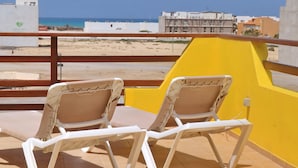 A balcony with two beige lounge chairs, a yellow wall, and a view of the beach and buildings in the background. - The Yellow Flat - Sea View (Boa Vista)